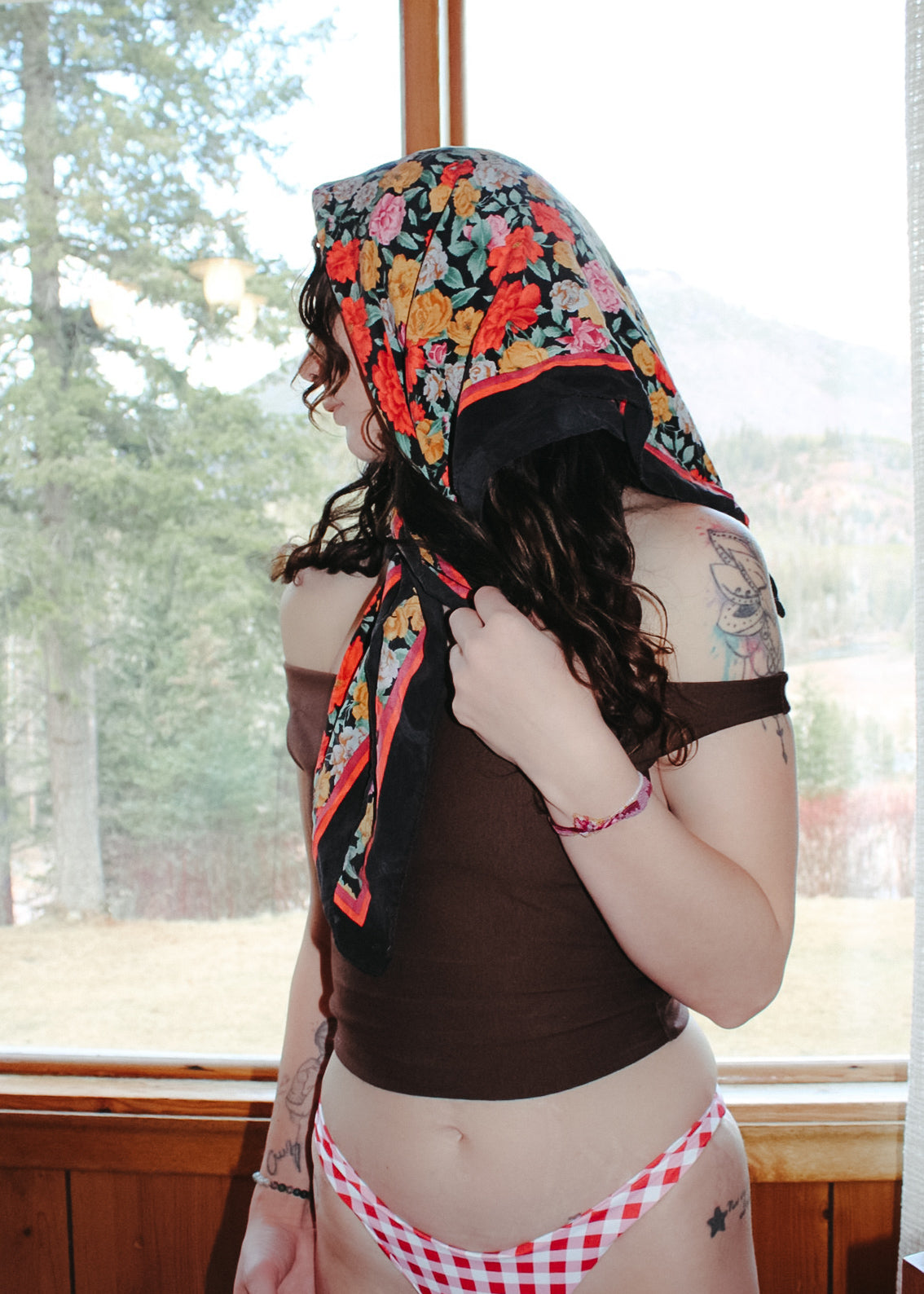Person wearing a floral headscarf and gingham bikini in front of a window with a view of trees.